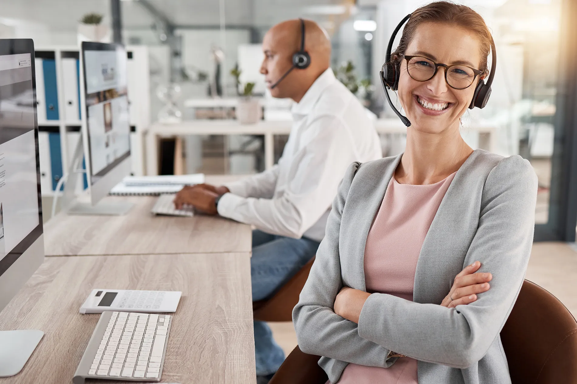 Mitarbeiterin mit Headset in einem modernen Kundenservice-Büro, unterstützt von einem Kollegen im Hintergrund.